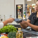 Cena ilustrativa de cartões e pagamentos: caixa de supermercado brasileiro com máquina de cartão genérica sem tela legível, cesta com alimentos e duas vias de comprovante em branco