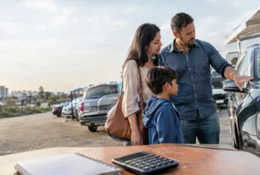 Cena ilustrativa de poupança e rendimentos: família brasileira olhando um carro usado em estacionamento aberto, com caderneta sem texto e calculadora sobre mesa em primeiro plano