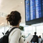 Cena ilustrativa de passagens aéreas e cartão: passageira em aeroporto brasileiro de costas olhando painel totalmente ilegível, segurando envelope com bilhetes sem escrita