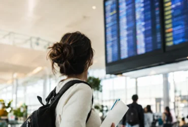Cena ilustrativa de passagens aéreas e cartão: passageira em aeroporto brasileiro de costas olhando painel totalmente ilegível, segurando envelope com bilhetes sem escrita