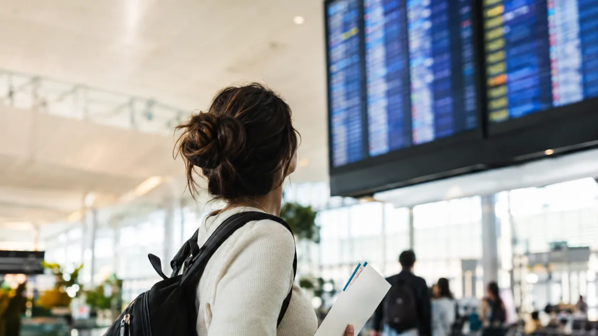 Cena ilustrativa de passagens aéreas e cartão: passageira em aeroporto brasileiro de costas olhando painel totalmente ilegível, segurando envelope com bilhetes sem escrita