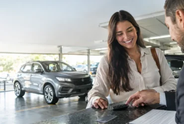 Cena ilustrativa de aluguel de veículos e cartão: carro alugado genérico em estacionamento iluminado, chave sobre balcão de locadora e cartão sem marca ao lado de contrato ilegível