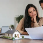Cena ilustrativa de financiamento e débito automático: casal observando boleto habitacional em branco diante de maquete pequena de casa, calendário desfocado e tela de banco apagada
