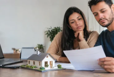 Cena ilustrativa de financiamento e débito automático: casal observando boleto habitacional em branco diante de maquete pequena de casa, calendário desfocado e tela de banco apagada