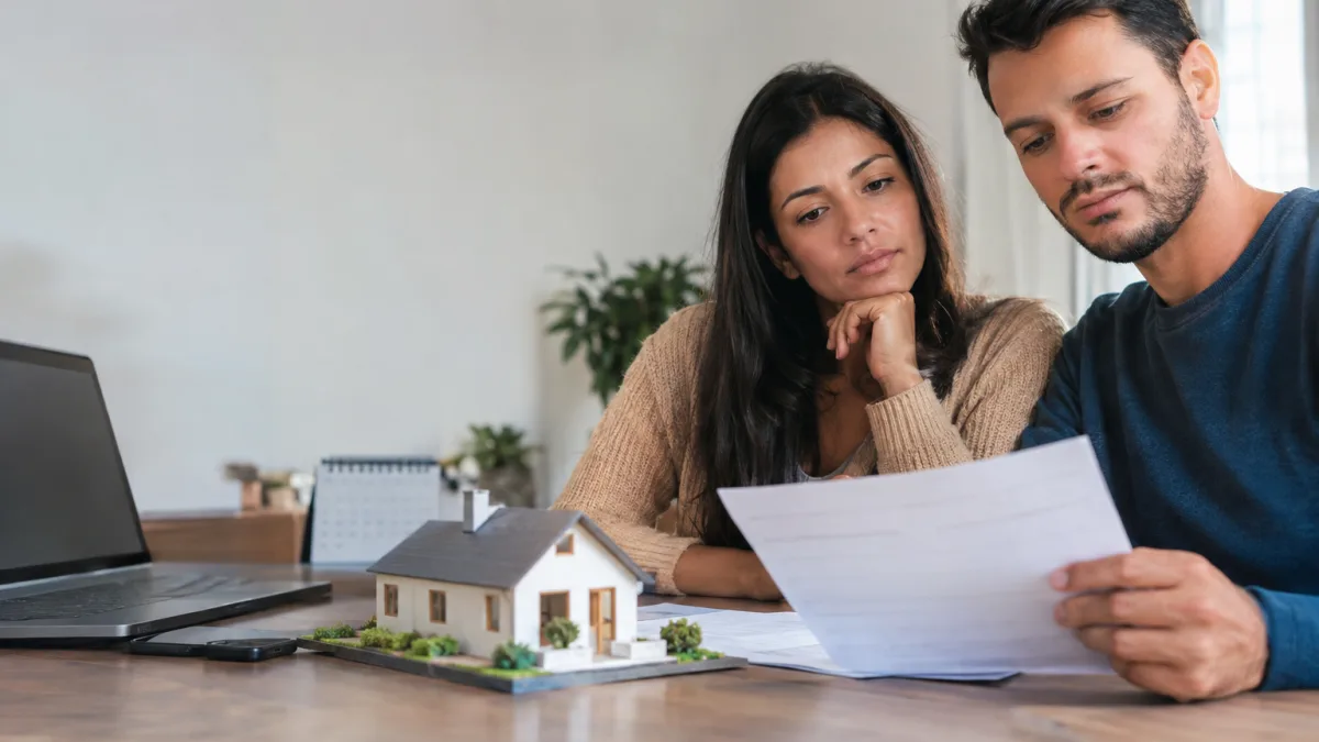 Cena ilustrativa de financiamento e débito automático: casal observando boleto habitacional em branco diante de maquete pequena de casa, calendário desfocado e tela de banco apagada