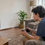 Cena ilustrativa de jogos digitais e cobranças: mãe e filho em sofá de casa brasileira, controle de videogame sem marca e cartão genérico sobre mesa, televisão sem conteúdo legível