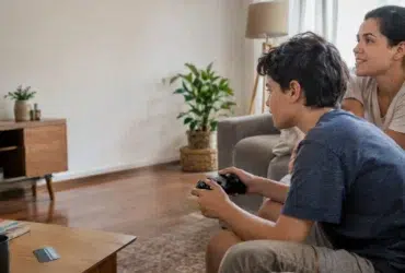 Cena ilustrativa de jogos digitais e cobranças: mãe e filho em sofá de casa brasileira, controle de videogame sem marca e cartão genérico sobre mesa, televisão sem conteúdo legível